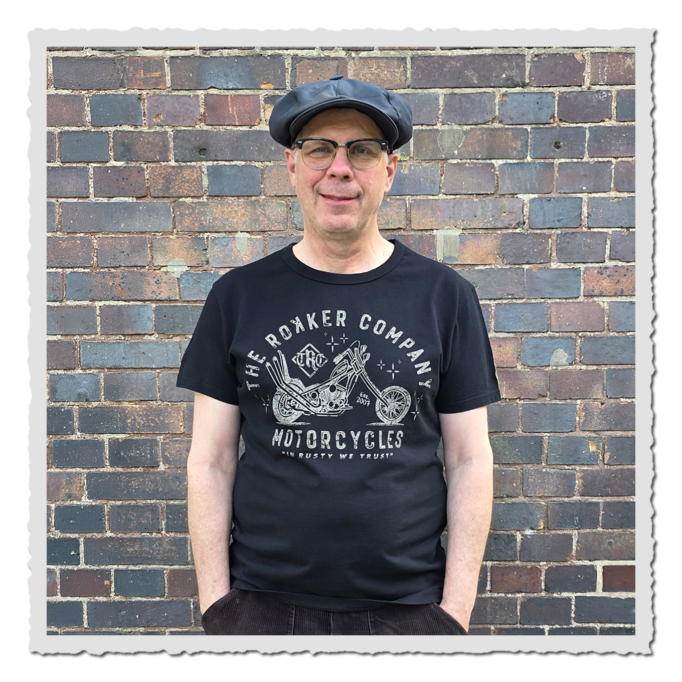 Mann von vorn in schwarzem T-Shirt mit hellem Motorradprint, dunkler Kappe, vor mehrfarbiger Backsteinwand.